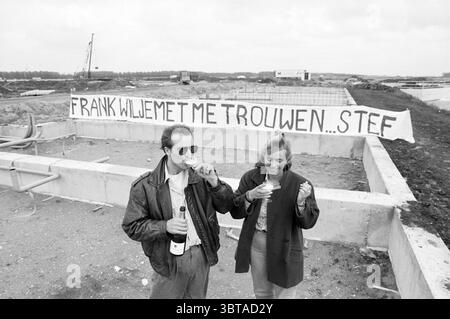 Banner Frank and Stef Velserbroek, Whizgle News, Dutch Desk, Niederlande, 1950 - 2000 am 18-06-1991. Diese Themen sind in der Abbildung dargestellt. In dieser Szene steht ein Paar in einer kargen, von unfertigen Bauarbeiten geprägten Umgebung im Freien. Der Boden darunter ist eine Mischung aus Schmutz und verstreuten Trümmern, was darauf hindeutet, dass sich das Gebiet in der frühen Phase der Entwicklung befindet. Der Mann, gekleidet in einer dunklen Lederjacke, scheint entspannt zu sein, während er lächelt, während er eine Flasche Champagner hält, was vielleicht einen Anlass feiert. Neben ihm trägt eine Frau ein helles Oberteil und hat ihr Haar mit Lose gestylt Stockfoto