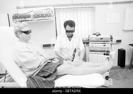 Physiotherapeut Mr. Feranci mit Lasergerät. Medizin Heilmethode, Whizgle News, Dutch Desk, Niederlande, 1950 - 2000 am 26-10-1987. Diese Themen werden in der Abbildung angezeigt. In einer klinischen Umgebung sitzt eine Frau auf einem Behandlungstisch und trägt eine dunkle Sonnenbrille, die einen Kontrast zu ihrem hellen Pullover bildet. Ihr kurzer Rock hat ein kariertes Muster, das der Szene eine gewisse Textur verleiht. Sie hat ein ruhiges Verhalten und wirkt trotz des medizinischen Kontextes entspannt. Neben ihr konzentriert sich ein Mann in einem weißen medizinischen Mantel aufmerksam auf seine Aufgabe. Er benutzt ein Gerät, um ihr Bein zu behandeln, das freigelegt ist, und enthüllt das c Stockfoto