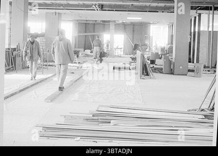 Collage Schiphol Schiphol, Whizgle News, Dutch Desk, Niederlande, 1950 - 2000 am 23-12-1970. Diese Themen sind in der Abbildung dargestellt. Die Szene zeigt eine Baustelle, die von einer Atmosphäre des Fleißes und der Transformation geprägt ist. Der Raum ist weitläufig, mit unfertigen Wänden und verschiedenen Materialien verstreut. Es sind mehrere Personen anwesend, die verschiedene Aufgaben wahrnehmen. Zwei Figuren bewegen sich auf einem überdachten Boden und scheinen sich auf ihre Aktivitäten konzentriert zu haben. Sie tragen praktische Arbeitskleidung, möglicherweise Jacken und Hosen, die der Szene ein utilitaristisches Gefühl verleihen. Die Indiv Stockfoto