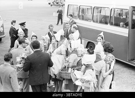 Cheese Girl Nelly Kraus bei Schiphol Cheese, Whizgle News, Dutch Desk, Niederlande, 1950 - 2000 am 02-10-1961. Diese Themen sind in der Abbildung dargestellt. Die Szene fängt ein lebhaftes Flughafengelände mit voller Aktivität und Spannung ein. Eine große Gruppe von Frauen steht vorne und in der Mitte, alle in passender Kleidung mit hellen gestreiften Kleidern und weißen Schürzen. Jede Frau trägt einen unverwechselbaren Kopfschmuck, möglicherweise einen bonnett oder einen ähnlichen Hut, der ihrem Aussehen einen Hauch von Gleichmäßigkeit und Charme verleiht. Sie halten bunte Blumensträuße, die sie dem Ankömmling gerne präsentieren wollen Stockfoto