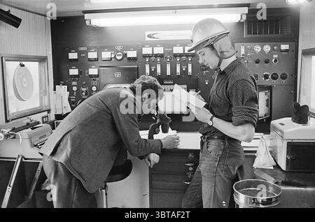 Rep. Oil Drilling Haarlemmermeer Report, Whizgle News, Dutch Desk, Niederlande, 1950 - 2000 am 10-02-1970. Diese Themen sind in der Abbildung dargestellt. Die Szene spielt in einem kompakten, industriellen Arbeitsbereich, der sich durch eine Vielzahl von Bedienfeldern und Instrumenten auszeichnet. Die Wände sind mit verschiedenen Anzeigen und Zifferblättern gesäumt, die von dunklen Farbtönen dominiert werden und eine utilitaristische Atmosphäre schaffen. Zwei Individuen besetzen den Vordergrund. Man untersucht einen kleinen Apparat und lehnt sich leicht nach vorne mit einem fokussierten Ausdruck. Diese Person trägt einen hellen Blazer, der sich von der lässigeren Kleidung des T abhebt Stockfoto