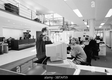 Neuer Schalter Zandvoort Zandvoort, Whizgle News, Dutch Desk, Niederlande, 1950 - 2000 am 03-12-1995. Dies sind die Elemente im Bild. Die Szene spielt in einer modernen Bürolobby mit einem geräumigen, luftigen Ambiente. Die Architektur ist mit großen Glasfenstern ausgestattet, die natürliches Licht in den Innenraum einströmen lassen und die Helligkeit insgesamt verbessern. Die Farbpalette besteht hauptsächlich aus weiß- und Hellgrau, was eine saubere und moderne Atmosphäre schafft. Vorne steht links ein Mann in dunkler Kleidung und interagiert mit einer Frau, die an einer Rezeption sitzt. Sie hat eine kurze blonde Ha Stockfoto