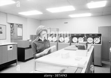 Frau am Honeywell 200 Computersystem (Punchkarten)., Whizgle News, Dutch Desk, Niederlande, 1950 - 2000. Diese Themen werden in der Abbildung angezeigt. Die Szene zeigt eine Frau, die in einer Retro-Büroumgebung steht, die ein Gefühl der Vergangenheit vermittelt, insbesondere der frühen Computerzeit. Sie hat ein poliertes Aussehen, mit gestylten Haaren und einer hübschen Bluse, die eine professionelle Umgebung andeutet. Im Hintergrund dominieren große, zylindrische Bandlaufwerke die Wand, deren metallische Oberflächen Licht reflektieren. Diese Geräte, die an die Technologie der 1960er und 1970er Jahre erinnern, sind in einer Reihe angeordnet, mit sichtbaren Rollen, die das sind Stockfoto