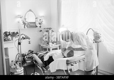 Maison Yvonne Beauty Institute Interior, Whizgle News, Dutch Desk, Niederlande, 1950 - 2000 am 23-11-1964. Dies sind die Elemente im Bild. Eine ruhige und intime Schönheitssalon-Szene entfaltet sich, in der sich der Patient bequem auf einem eleganten, modernen Behandlungsstuhl zurücklehnen kann. Die Person scheint eine Gesichtsbehandlung zu genießen, indem sie sich mit einem Handtuch zurücklehnt, das sanft über die Schultern gelegt wird. Im Vordergrund konzentriert sich eine Kosmetikerin in einer knackig weißen Uniform auf ihre Aufgabe und zeigt Professionalität und Sorgfalt. Sie ist mit einem Hauch von Ruhe konfrontiert, ihr Verhalten vermittelt ein Gefühl von Vertrauen und Re Stockfoto