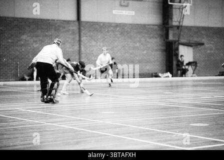Erdbeeren - HBS Hockey Hallen, Whizgle News, Dutch Desk, Niederlande, 1950 - 2000 am 14-02-1983. Diese Themen sind in der Abbildung dargestellt. Die Szene zeigt eine dynamische Sportszene, wahrscheinlich ein Gymnasium. Der Boden aus poliertem Holz ist mit weißen Linien markiert, die verschiedene Abschnitte für das Gameplay abgrenzen. Im Vordergrund hockt ein Torhüter, der in Schutzkleidung gekleidet ist, tief in die Hocke, bereit zu reagieren. Ihre Aufmerksamkeit richtet sich auf einen Spieler, der auf ihn zusprintet, der in der Hand bleibt, während er sich darauf vorbereitet, den Ball zu schlagen. Die Kleidung des Torhüters steht in starkem Kontrast zu den dunklen Leggings und dem T-Shirt Stockfoto