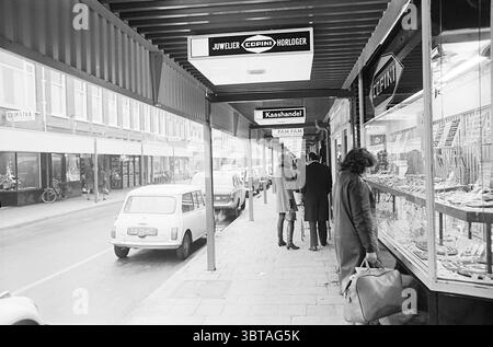 Überblick über die Cronjéstraat mit Baldachinen Cronjestraat Haarlem Generaal Cronjéstraat Niederlande, Whizgle News, Dutch Desk, Niederlande, 1950 - 2000 am 12-12-1969. Das Bild enthält diese Themen. Die Szene zeigt eine geschäftige Stadtstraße mit Schwerpunkt auf einer Reihe von Geschäften unter einem überdachten Gang. Die Architektur verfügt über eine Markise in gedämpftem Ton, die sanfte Schatten entlang des Gehwegs wirft. Auf der linken Seite zeigen verschiedene Displays eine Reihe von Waren, die unter hellem Licht glitzern. Dekorative Gegenstände und Schmuck sind sorgfältig angeordnet und betonen ihre komplexen Designs. Das g Stockfoto
