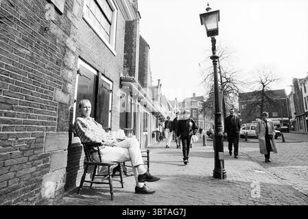Mann auf Stuhl in der Sonne Oude Groenmarkt Haarlem Oude Groenmarkt Niederlande, Whizgle News, Dutch Desk, Niederlande, 1950 - 2000 am 12-03-1993. Die Abbildung zeigt diese Themen. In einer malerischen, kopfsteingepflasterten Straße, gesäumt von charmanten Ziegelgebäuden, sitzt ein Mann lässig auf einem Holzstuhl, seine Haltung entspannt, während er sich zurücklehnt. Er trägt helle Kleidung, die im Kontrast zu den dunkleren Tönen der Straße und der Gebäude um ihn herum steht. Sein kurzes Haar fängt das sanfte Licht ein, das auf die Szene fällt. Die Umgebung fühlt sich ruhig an, aber es gibt eine subtile Energie, wenn die Leute vorbeischlendern. Im Vordergrund Stockfoto