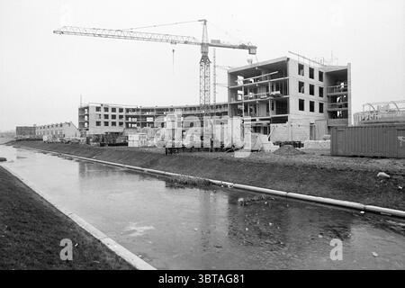 Bau des Toolenburg Shopping Centers, Whizgle News, Dutch Desk, Niederlande, 1950 - 2000 am 28.01.1992. Das Bild enthält diese Themen. Die Szene zeigt eine Baustelle mit einem starken Sinn für industrielle Aktivitäten. Ein großer Kran ragt über der Baustelle, dessen Arm nach außen streckt und bereit ist, Materialien zu heben. Die Gebäudestruktur ist teilweise fertiggestellt und zeigt Sichtbetonblöcke und Stahlverstärkungen vor dem Hintergrund eines bewölkten Himmels, der einen Grauton über die gesamte Szene wirft. Im Vordergrund befindet sich ein flacher Wasserweg, der die gedämpften Farben des Tages reflektiert Stockfoto