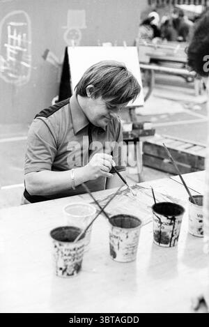 Stuif - Stuif für Behinderte Stuif, Whizgle News, Dutch Desk, Niederlande, 1950 - 2000 am 06-08-1977. Diese Themen sind in der Abbildung dargestellt. Die Szene fängt ein kreatives Umfeld ein, das mit Freude und Konzentration durchdrungen ist. Im Vordergrund sitzt eine junge Person an einem Holztisch, ihre Haltung reflektiert die Konzentration, während sie einen Pinsel in einen Topf mit leuchtender Farbe taucht. Die Kleidung ist lässig und farbenfroh, mit einem Hemd, das auf eine verspielte, aber dennoch ansprechende Art und Weise für künstlerische Unternehmungen hinweist. Um den Tisch herum befinden sich mehrere Behälter mit verschiedenen Farbtönen, deren Oberflächen mit einem gekennzeichnet sind Stockfoto