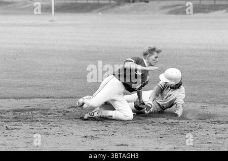 Softball Sparks - Bloemendaal, Whizgle News, Dutch Desk, Niederlande, 1950 - 2000 am 15-06-1991. Das Bild enthält diese Themen. In einer dynamischen Sportszene spielen zwei junge Athleten einen entscheidenden Moment auf einem Baseballdiamanten. Der erste Athlet, der an der Spitze steht, befindet sich in der Mitte der Action und rutscht mit einem entschlossenen Ausdruck in die Basis. In einer kontrastierenden Uniform – einem leichteren Hemd mit auffälligen Schriftzügen und einer dunklen Hose – wird seine Form in einer dramatischen, sportlichen Pose festgehalten. Staub und Schmutz werden um ihn herum geschleudert, was die Intensität seiner Bemühungen unterstreicht. Der zweite Athlet, bereit Stockfoto