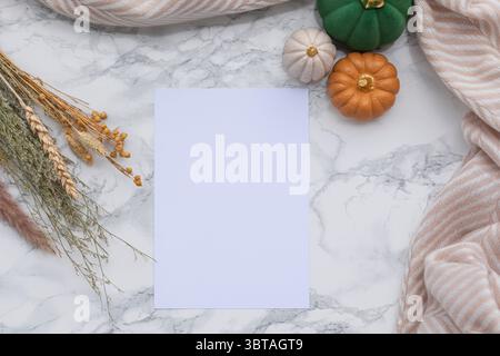 Gestylte, flache Herbstlage mit leerem weißem Papier, umgeben von einem beigefarbenen Schal, getrockneten Pflanzenstoffen, Samtkürbissen auf Marmoroberfläche, perfekt für saisonale Werbung Stockfoto