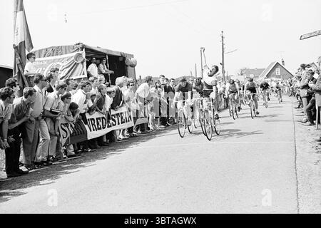 Cycling Race., Whizgle News, Dutch Desk, Niederlande, 1950 - 2000. Das Bild enthält diese Themen. In einer lebhaften Outdoor-Szene findet ein temperamentvolles Radrennen statt. Die Fahrbahn ist gesäumt von einer Menge begeisterter Zuschauer, deren Gesichter vor Aufregung und Ermutigung animiert sind, während sie die Teilnehmer anfeuern. Die Atmosphäre strahlt ein Gefühl der Gemeinschaft und des Feierns aus. Die Zuschauer tragen legere Kleidung aus der Mitte des 20. Jahrhunderts – einige tragen leichte Hemden und Shorts, während andere Hüte tragen, um sich vor der Sonne zu schützen. Die in markanten Trikots gekleideten Radfahrer sind prominent positioniert Stockfoto