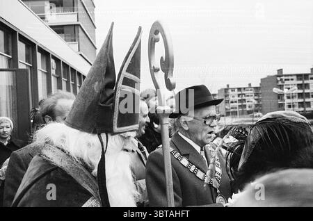 Ankunft von St. Nicholas in Beverwijk St. Nicholas Sinterklaas, Whizgle News, Dutch Desk, Niederlande, 1950 - 2000 am 16. 11. 1974. Dies sind die Elemente im Bild. Die Szene zeigt eine Zusammenkunft von Einzelpersonen in einer Umgebung im Freien, die durch eine soziale oder festliche Atmosphäre gekennzeichnet ist. Im Vordergrund fallen zwei prominente Figuren auf: Eine trägt ein kunstvolles Kostüm, das dem Heiligen Nikolaus ähnelt, komplett mit einem königlichen umhang und einem hohen, spitzen Hut, der mit einem großen, dekorativen Emblem verziert ist. Sein langer weißer Bart trägt zur traditionellen Erscheinung bei. Die andere Figur, gekleidet in einen formellen Anzug und Stockfoto