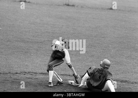 Softball Pioneers - HVV, Whizgle News, Dutch Desk, Niederlande, 1950 - 2000 am 08-06-1991. Diese Themen sind in der Abbildung dargestellt. In einer dynamischen Szene auf einem Softballfeld spielen zwei Spieler einen entscheidenden Moment. Ein Athlet in einem weiß-tiefblauen Trikot mit passender Hose scheint in Bewegung zu sein, nachdem er gerade die Basis markiert hat. Sie haben dunkles, gewelltes Haar, das hinter ihnen fließt und ein Gefühl von Dringlichkeit und Handlung vermittelt. Der andere Spieler in einem dunkelfarbenen Trikot mit der Nummer 4 liegt auf dem Boden und scheint gerade die Basis erreicht zu haben. Sie trägt einen Helm, der rot erscheint, im Kontrast zu wi Stockfoto