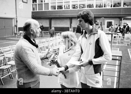 Tennis Awards Zeremonie, Whizgle News, Dutch Desk, Niederlande, 1950 - 2000 im Jahr 1980. Dies sind die Themen im Bild. Die Szene fängt einen Moment der Interaktion in einer Sporthalle ein, vermutlich während einer Veranstaltung oder Feier. Im Vordergrund stehen zwei junge Individuen, ein Mann und eine Frau, gegenüber einem älteren Herrn. Der jüngere Mann, gekleidet in einem weißen sportlichen Trainingsanzug mit dezenten blauen Akzenten, hält ein Stück Papier in der einen Hand, während er die andere ausdehnt, um einen Blumenstrauß von dem älteren Mann zu erhalten, der in einem strukturierten Strickpullover verziert ist. Die junge Frau, die einen trägt Stockfoto