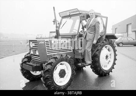 Herr Segers Agrotechniek Noordwijkerhout Persons Companies Noordwijkerhout, Whizgle News, Dutch Desk, Niederlande, 1950 - 2000 am 16-11-1984. Dies sind die Themen im Bild. Ein Mann in einem Anzug sitzt souverän auf einem robusten Traktor mit auffälligen Schwarz-weiß-Details. Die großen, robusten Reifen des Traktors verfügen über tiefe Rillen, die das robuste Design unterstreichen. Die Umgebung scheint ein Außenbereich zu sein, möglicherweise ein Bauernhof oder ein landwirtschaftlicher Ausstellungsraum, umgeben von einer grauen Atmosphäre, die auf einen bewölkten Himmel oder ein nebeliges Wetter hindeutet. Im Hintergrund befindet sich ein großes Gebäude, möglicherweise ein Stockfoto