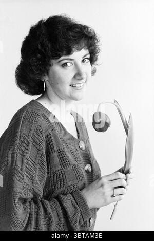 American Laura A@ron im Frans Hals Museum Haarlem Groot Heiligland Niederlande, Whizgle News, Dutch Desk, Niederlande, 1950 - 2000 am 09-04-1992. Das Bild enthält diese Themen. In dieser Szene wird eine Person mit lockigem, dunklem Haar so positioniert, dass ihre Merkmale und ihr Ausdruck hervorgehoben werden. Sie tragen einen strukturierten Pullover mit einem Muster aus Quadraten, das durch sein kompliziertes Design visuell interessant ist. Die dezente Farbe des Pullovers trägt zu einem ruhigen Ambiente bei. Der einzelne hält eine einzelne Tulpe, die sich deutlich von den gedämpften Tönen des Surroundi abhebt Stockfoto