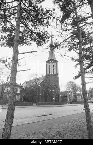 Äußere der St. Gregor Kirche in IJmuiden Churches, Whizgle News, Dutch Desk, Niederlande, 1950 - 2000 am 16-01-1977. Die Abbildung zeigt diese Themen. Die Szene zeigt eine markante Kirche, die sich durch einen hohen Turm und einen markanten Uhrenturm auszeichnet. Das Gebäude ist in einem traditionellen architektonischen Stil gebaut, mit einer dunklen Ziegelfassade, die einen subtilen Kontrast zu den sanfteren Tönen der Umgebung bildet. Zwei hohe, schlanke Bäume säumen die Kirche im Vordergrund, ihre Rinde strukturiert und rau, mit Ästen, die sich ausbreiten und zarte Schatten bilden. Die Bäume bilden eine natürliche Grenze Stockfoto