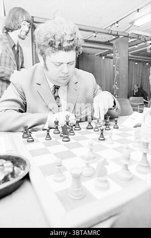 Eröffnung der vier-Wege-Veranstaltung Blast Furnace Chess Tournament, Whizgle News, Dutch Desk, Niederlande, 1950 - 2000 am 15.01.1972. Das Bild enthält diese Themen. In einem belebten Innenbereich sitzt ein fokussierter Mann an einem Schachtisch, der tief in ein Match verwickelt ist. Er trägt einen strukturierten, hellen Anzug, der den satten, dunklen Farbtönen der hölzernen Schachfiguren auf dem Brett vor ihm gegenübersteht. Sein Haar ist lockig und voluminös, was seinem Aussehen einen Vintage-Charme verleiht. Er beugt sich leicht nach vorne, seine Hand steht über einen schwarzen Bauern und denkt über seinen nächsten Zug nach. Das Schachbrett selbst ist ein klassisches Karomuster Stockfoto