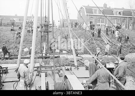 1st Pile Bootshaus First Pile, Whizgle News, Dutch Desk, Niederlande, 1950 - 2000 am 06-05-1968. Das Bild enthält diese Themen. Die Szene fängt eine geschäftige Baustelle mit einem Gewässer ein, das durch eine Mischung aus Aktivität und Beobachtung gekennzeichnet ist. Im Vordergrund arbeitet eine Gruppe von Arbeitern in praktischer, zweckmäßiger Kleidung mit schweren Maschinen, die von großen Zahnrädern und Riemenscheiben dominiert werden. Ihre Gesichter zeigen Konzentration, während sie die Ausrüstung verwalten. Um sie herum versammelt sich eine kleine Menschenmenge, die mit Neugier das Geschehen beobachtet. Einige Personen erscheinen Stockfoto