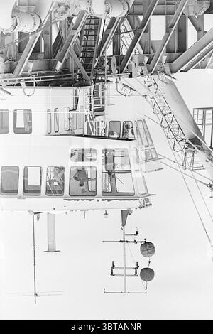 Sandsauger., Whizgle News, Dutch Desk, Niederlande, 1950 - 2000. Diese Themen werden in der Abbildung angezeigt. Die Szene erfasst eine komplexe industrielle Struktur, die überwiegend mit einem großen, metallischen Rahmen versehen ist. Die Elemente der Struktur sind in Grautönen gestrichen und stehen im Kontrast zur hellen Umgebung. Hervorzuheben ist eine in einem dramatischen Winkel angeordnete Kontrollkabine mit großen Glasfenstern, die eine Sicht in den Innenraum ermöglichen. In der Kabine sind zwei Personen sichtbar, deren Merkmale verdeckt sind, aber auf Konzentration hinweisen, da sie mit der Maschine in Berührung kommen. Stockfoto