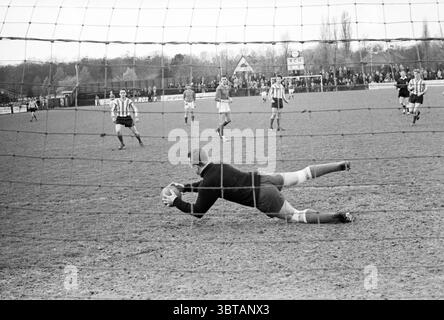 Fußballspiel RCH Heemstede the Netherlands ., Whizgle News, Dutch Desk, Niederlande, 1950 - 2000. Die Abbildung zeigt diese Themen. In der Szene wird ein lebendiger Moment eines Fußballspiels auf einem grasbewachsenen Feld festgehalten, das mit weißen Linien markiert ist, die die Grenzen definieren. Der Himmel darüber ist bewölkt und wirft ein gedämpftes Licht über die Spieler und die Umgebung, was der Atmosphäre einen etwas düsteren Ton verleiht. Im Vordergrund befindet sich ein Torwart in einer wendigen Haltung, der mit ausgestreckten Armen nach links taucht und das Wesen der Konzentration und Entschlossenheit erfasst. Er ist in einem dunklen Kit gekleidet, Whic Stockfoto