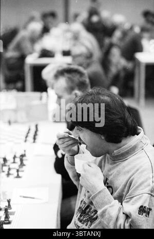 Blastovens Chess Tournament Wijk aan Zee, Whizgle News, Dutch Desk, Niederlande, 1950 - 2000 am 19.01.1993. Das Bild enthält diese Themen. Die Szene spielt in einem geräumigen Innenbereich, wahrscheinlich in einer Turnierhalle, mit langen Tischen, die in einem ordentlichen Raster angeordnet sind. Auf diesen Tischen sind Schachbretter sorgfältig angelegt, die jeweils mit Spielstücken versehen sind. Die Atmosphäre ist komponiert und konzentriert, mit einer Mischung aus Intensität und Gelassenheit unter den Teilnehmern. Im Vordergrund sitzt eine Person an einem Tisch, spielt in einem Schachspiel und isst gleichzeitig aus einer kleinen Schüssel. Sie tragen eine LIG Stockfoto