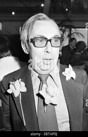 Präsentation Blumenmädchen Holz und öffentliche Gärten Service Blumen und Blumenmädchen öffentliche Gärten Pflanzen Sträucher Heister, Whizgle News, Dutch Desk, Niederlande, 1950 - 2000 am 29-03-1977. Diese Themen sind in der Abbildung dargestellt. Die Szene zeigt eine Person mit einem ausdrucksstarken Auftreten, die in der Mitte steht. Er hat silbergraue Haare, die ordentlich gestylt sind, und trägt eine große, dunkle Brille, die ihm ein außergewöhnliches, durchdachtes Aussehen verleiht. Sein Ensemble besteht aus einem dunklen Anzug, dessen Revers mit zwei leuchtenden Narzissen verziert ist, die sich von den düsteren Farben seiner Kleidung abheben und einen Hauch von Glanz verleihen Stockfoto