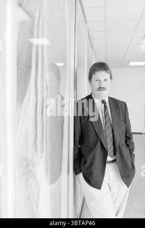 Mari van der Vlis Sprecherin für Hoogovens, Whizgle News, Dutch Desk, Niederlande, 1950 - 2000 am 17.06.1992. Diese Themen sind in der Abbildung dargestellt. In einem gut beleuchteten Innenraum steht ein Mann mittleren Alters selbstbewusst neben einer Glaswand, die sein Bild widerspiegelt. Er hat einen hübschen Schnurrbart und trägt ein formelles Outfit, bestehend aus einem dunklen Blazer und einem hellen Hemd, ergänzt durch eine gemusterte Krawatte, die einen Hauch von visuellem Interesse verleiht. Die hellen Farben seines Anzugs stehen im Kontrast zum tiefen Farbton seines Blazers. Das Ambiente hat eine moderne, minimalistische Ästhetik, mit klaren Linien und einem geräumigen Raum Stockfoto