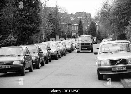Traffic Platanenlaan Bloemendaal Platanenlaan, Whizgle News, Dutch Desk, Niederlande, 1950 - 2000 am 07-04-1991. Dies sind die Themen im Bild. Die Szene zeigt eine enge Straße, die von geparkten Autos auf beiden Seiten gesäumt ist und einen engen Weg bildet. Die Fahrzeuge, meist kompakt und etwas veraltet, sind in ordentlichen Reihen angeordnet und zeigen eine Vielzahl von Farben, darunter Silber, Dunkelgrün und Rot, mit einigen Anzeichen von Verschleiß. Im Vordergrund fährt ein größeres, kastenförmiges Fahrzeug die Mitte der Straße hinunter, etwas betont durch seine Position vor dem Hintergrund des geparkten c Stockfoto