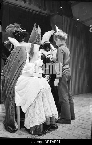 Sinterklaas Party im Wijsmuller Wijsmuller, Whizgle News, Dutch Desk, Niederlande, 1950 - 2000 am 06.12.1969. Dies sind die Elemente im Bild. Die Szene fängt einen festlichen Moment ein, wahrscheinlich während einer Feier oder Veranstaltung, die sich an Kinder richtet. Im Vordergrund sitzt eine Frau in einem langen, weißen Kleid mit komplizierten Spitzendetails bequem auf einem Stuhl. Sie trägt einen auffälligen, hohen, rot-weißen Kopfschmuck, der ihrem Aussehen einen königlichen Ton verleiht. Ihre Gesichtszüge sind teilweise verdeckt, was einen Hauch von Mystik hinzufügt. Um sie herum sind lebendige Figuren, die wahrscheinlich festliche Charaktere darstellen Stockfoto
