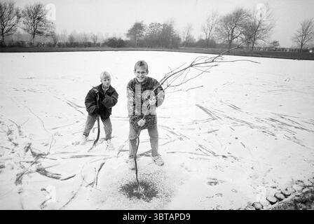 Ice Fun Santpoort Santpoort, Whizgle News, Dutch Desk, Niederlande, 1950 - 2000 am 21-11-1993. Diese Themen sind in der Abbildung dargestellt. In einer ruhigen Winterlandschaft spielen zwei Kinder auf einem verschneiten Feld, das sich vor ihnen ausdehnt. Der Boden ist mit einer weichen, weißen Schneedecke bedeckt, die unter gedämpftem Tageslicht glitzert. Der Himmel ist bedeckt und wirft einen grauen Farbton über die Szene, was die winterliche Atmosphäre verstärkt. Ein Kind, das im Vordergrund steht, hält selbstbewusst mehrere lange Zweige, die er gesammelt hat, in der Hand, als wäre es bereit, aus seinem Fund etwas zu erschaffen. Er trägt einen Patte Stockfoto