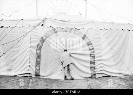 Circus Renz Schalkwijk Schalkwijk, Whizgle News, Dutch Desk, Niederlande, 1950 - 2000 am 08-05-1990. Diese Themen werden in der Abbildung angezeigt. In einer monochromatischen Szene zeichnet sich ein großes, rundes Zelt durch seine besondere Architektur aus, mit einem anmutig gewölbten Eingang, der von Sternen flankiert wird. Der Stoff des Zeltes scheint leicht abgenutzt zu sein, was darauf hindeutet, dass es viele Ereignisse erlebt hat. Eine dezente Textur ist sichtbar, die auf die Robustheit des Materials und den Verschleiß durch Außenbedingungen hinweist. In der Nähe des Eingangs ist eine Einzelfigur abgebildet, die teilweise verdeckt ist, wenn sie sich hineinlehnen, um hineinzuschauen. Die Person ist verkleidet Stockfoto
