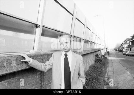Herr Dorst bei Lärmschutzvorrichtungen Randweg Lärmschutzpersonen Westelijke Randweg, Whizgle News, Dutch Desk, Niederlande, 1950 - 2000 unter 30-12-1983. Diese Themen werden in der Abbildung angezeigt. In einer monochromen Szene steht ein Mann in der Nähe einer niedrigen Steinmauer und lehnt sich leicht gegen sie. Er trägt einen hellen Anzug mit dunkler Krawatte, der einen auffälligen Kontrast zum gedämpften Hintergrund bildet. Sein Ausdruck wirkt kontemplativ, da er leicht zur Seite blickt und zu einer reflektierenden Stimmung beiträgt. Hinter ihm erhebt sich eine transparente Barriere aus Paneelen, die eine faszinierende visuelle Linie schafft, die das Auge nach oben zieht Stockfoto