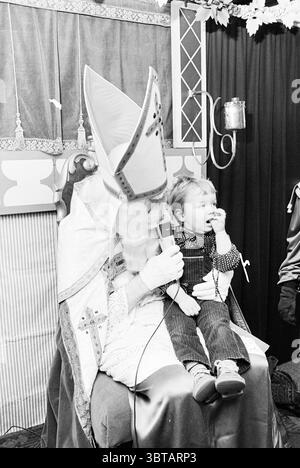 Sinterklaas on HD St. Nicholas Sinterklaas, Whizgle News, Dutch Desk, Niederlande, 1950 - 2000 am 28-11-1979. Diese Themen sind in der Abbildung dargestellt. In einem lebhaften Innenbereich steht eine Figur in einem raffinierten, traditionellen Outfit im Vordergrund. Diese Figur ist mit einem hohen, spitzen Hut mit verzierten Mustern und einem langen Gewand mit hellen Farben und Verzierungen verziert. Das Outfit vermittelt ein Gefühl von Autorität und Festlichkeit, was auf eine kulturelle oder zeremonielle Bedeutung hindeutet. Die Figur sitzt bequem in einem großen Stuhl und hält ein Mikrofon, was auf eine Interaktion mit dem hindeutet Stockfoto