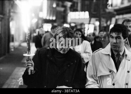 Vorbei an der Fackel Haarlem City of Light Haarlem the Netherlands, Whizgle News, Dutch Desk, Niederlande, 1950 - 2000 am 01-05-1991. Das Bild enthält diese Themen. In einer geschäftigen städtischen Umgebung bei Nacht wird eine vielfältige Gruppe von Menschen bei ihren abendlichen Aktivitäten gefangen genommen. Die Szene wird durch eine Mischung aus künstlichem Licht beleuchtet, das ein warmes Leuchten vor der kühlen, dunklen Kulisse der Straßen ausstrahlt. Im Vordergrund steht eine Frau, die einen leuchtenden Funkler hält. Sie hat kurze Haare und trägt eine große Brille, ihr Ausdruck besinnlich, während sie nach vorne blickt. In einem dunklen Mantel gekleidet, bildet sie einen scharfen Kontrast Stockfoto