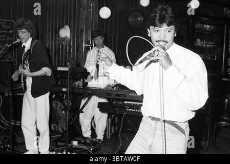 Zijlpoort singendes Festival Gesang und Gesangschöre Haarlem the Netherlands, Whizgle News, Dutch Desk, Niederlande, 1950 - 2000 am 29-04-1989. Das Bild enthält diese Themen. In einem lebendigen Indoor-Setting treten drei Musiker auf der Bühne auf, die Energie und Leidenschaft ausstrahlen. Das Ambiente ist mit einer warmen, aber dennoch gedämpften Beleuchtung erfüllt, die ihre Merkmale hervorhebt und sanfte Schatten um sie herum wirft. Der erste Darsteller, der an der Vorderseite deutlich positioniert ist, hat eine selbstbewusste Haltung, während er ein Mikrofon hält. Er hat dunkles Haar, mit einer leichten Welle gestylt und ist in einem knusprigen weißen Hemd gekleidet, das sich an den Tag legt Stockfoto