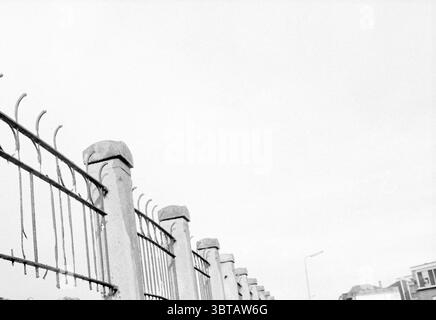Bad Walls Bik and Arnoldkade IJm IJmuiden the Netherlands, Whizgle News, Dutch Desk, Niederlande, 1950 - 2000 am 22-04-1991. Dies sind die Elemente im Bild. Die Szene zeigt einen langen, verwitterten Zaun, der sich über den Vordergrund erstreckt und mit scharfem, verdrehtem Stacheldraht gekrönt ist. Die Zaunpfähle sind aus Zement und weisen eine raue Textur auf, die Anzeichen von Verschleiß und Alter mit sichtbaren Rissen und Unregelmäßigkeiten zeigt. Die Gesamtfarbpalette ist monochromatisch, dominiert von verschiedenen Grautönen, was der Komposition ein schlichtes, düsteres Gefühl verleiht. Das Licht scheint gestreut zu sein, was auf einen bedeckten Himmel hindeutet Stockfoto