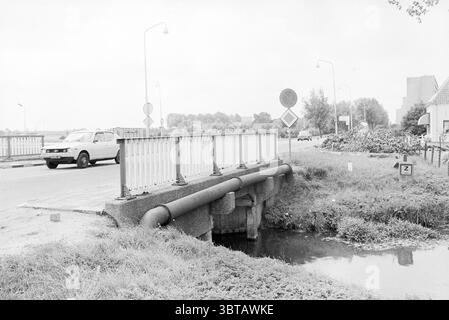 Brücke in der ländlichen Region Heemskerk, Whizgle News, Dutch Desk, Niederlande, 1950 - 2000 am 09-07-1979. Das Bild enthält diese Themen. Die Szene präsentiert eine ruhige ländliche Umgebung, dominiert von einer einfachen Brücke, die über einen schmalen Wasserweg führt. Die Brücke verfügt über eine stabile Betonkonstruktion mit einer niedrigen Barriere aus Metallgeländern, die weiß lackiert ist, aber Anzeichen von Verschleiß aufweist. Die Straße, die zur Brücke führt, ist sichtbar, mit Markierungsschildern, die die Verkehrsvorschriften in der Nähe anzeigen. Rund um die Brücke ist die Landschaft vorwiegend grün, geprägt von üppig und gut anmutendem Gras. Stockfoto