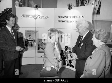 Verleihung einer hohen königlichen Auszeichnung an einen Beamten des Arbeitsamtes, Whizgle News, Dutch Desk, Niederlande, 1950 - 2000 im Jahr 1992. Das Bild enthält diese Themen. In einem gut beleuchteten Innenbereich gibt es eine Gruppe von Einzelpersonen, die sich in der Nähe einer Kulisse aus mehreren Displays an einem Gespräch beteiligen. Die Kulisse bietet eine Reihe von Panels mit verschiedenen Bildern und grafischen Elementen, die eine informative Atmosphäre schaffen. Die Farben neigen überwiegend zu monochromen Tönen mit Elementen in weiß, Grau und Schwarz. Im Vordergrund ein Mann mit leicht lockigen Haaren und einer gepflegten App Stockfoto