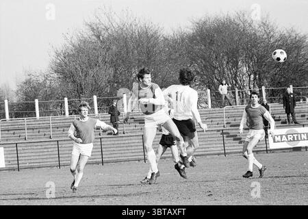 B.M.H.C? - Victoria Hockey, Whizgle News, Dutch Desk, Niederlande, 1950 - 2000 am 28.02.1971. Das Bild enthält diese Themen. Die Szene fängt einen intensiven Moment auf einem Sportplatz ein, der auf ein Fußballspiel hinweist. Im Vordergrund stehen zwei Spieler – einer in der Luft, der springt, um den Ball zu steuern, der direkt über ihm hängt. Er trägt ein helles Hemd, während ein rot-weißes Trikot auf einem anderen Spieler zu sehen ist, der sich in der Nähe befindet und die Action beobachtet. Um sie herum zeigen zusätzliche Spieler in ähnlichen Uniformen eine Reihe von expre Stockfoto