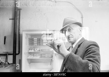 Report Tobacco Plantage Tobacco, Whizgle News, Dutch Desk, Niederlande, 1950 - 2000 am 11-06-1968. Die Abbildung zeigt diese Themen. Die Szene zeigt einen älteren Mann in einem schwach beleuchteten, leicht übersichtlichen Innenraum. Er trägt einen dunklen, strukturierten Anzug, begleitet von einem stilvollen Filzhut, der seinem Aussehen einen Hauch von Raffinesse verleiht. Sein Ausdruck wirkt nachdenklich, und er hält ein kleines Objekt – möglicherweise einen Kanister oder Behälter – dicht vor sich und lenkt die Aufmerksamkeit darauf. Im Hintergrund befindet sich ein offener Kühlschrank, in dem Regale mit verschiedenen Gegenständen, von denen einige scheinbar zu sein scheinen, sichtbar werden Stockfoto