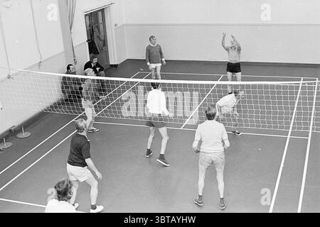Volley-Ball., Whizgle News, Dutch Desk, Niederlande, 1950 - 2000. Dies sind die Elemente im Bild. Die Szene fängt ein Indoor-Volleyballspiel ein, das durch eine energiegeladene und lebhafte Atmosphäre gekennzeichnet ist. Der Raum verfügt über einen glatten, polierten Holzboden, der die Deckenleuchten reflektiert, die eine helle Beleuchtung im Raum erzeugen. Im Vordergrund nimmt eine Gruppe von sechs Personen aktiv am Spiel Teil. Sie tragen legere Sportkleidung, einige tragen Shorts und kurzärmelige Hemden, andere tragen Langarm. Die Farbpalette umfasst eine Mischung aus lebhaften und neutralen Tönen Stockfoto