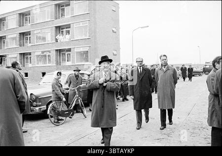 Bürgermeister gibt 1. Schlüsselübergabe Schalkwijk und überträgt Wohnungsbau, Whizgle News, Dutch Desk, Niederlande, 1950 - 2000 am 27.02.1961. Das Bild enthält diese Themen. Die Szene spielt in einer urbanen Umgebung, die wahrscheinlich aus der Mitte des 20. Jahrhunderts stammt und von einer Kulisse aus bescheidenen, mehrstöckigen Backsteinbauten geprägt ist. Die Architektur hat ein utilitaristisches Gefühl, mit großen Fenstern, die Blicke auf Menschen freigeben, die hinausschauen, was ein Gefühl von Gemeinschaft oder Neugier suggeriert. Im Vordergrund läuft eine Gruppe von Männern auf einem breiten Betonweg. Der Mann vorne trägt einen dunklen Mantel und einen Stockfoto