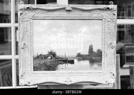 Fa. Artistic The Bewunderung IJmuiden IJmuiden the Netherlands, Whizgle News, Dutch Desk, Niederlande, 1950 - 2000 am 09-04-1995. Die Abbildung zeigt diese Themen. In einer ruhigen Umgebung umgibt ein wunderschön gefertigter Rahmen eine ruhige Landschaftsszene. Der Rahmen ist verziert und mit aufwändigen Schnitzereien versehen, die einen Hauch von Eleganz verleihen. Innerhalb dieses Rahmens fängt die gemalte Szene ein friedliches Flussbett unter einem weichen, bewölkten Himmel ein. Das Wasser im Fluss reflektiert die gedämpften Farben des Himmels und schafft ein Gefühl der Harmonie. Links befindet sich eine rustikale Struktur aus Holz, teilweise hidd Stockfoto