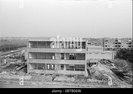 Haushaltsschule Delftlaan Gebäude Haarlem Delftlaan Niederlande, Whizgle News, Dutch Desk, Niederlande, 1950 - 2000 am 15-10-1963. Die Abbildung zeigt diese Themen. Die Szene präsentiert ein teilweise gebautes modernes Gebäude, das sich durch klare Linien und große Glasfenster auszeichnet. Die Struktur hat eine schlanke, metallische Fassade, die eine dezente Palette von Grau und Silber reflektiert und sich in den bewölkten Himmel einfügt. Um das Gebäude herum schaffen Baumaterialien wie Holzdielen und Metallgerüste eine Atmosphäre der ständigen Entwicklung. Im Vordergrund ist die Erde gestört Stockfoto