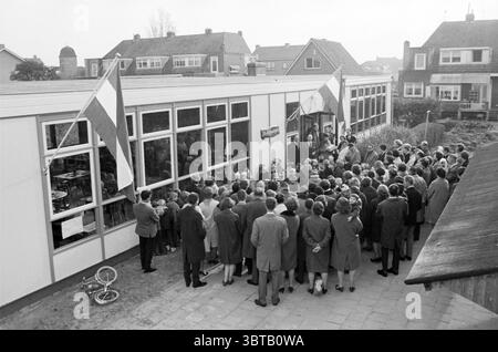 Eröffnung der Schule de Wilgenkatjes Zwanenburg Wilgenlaan., Whizgle News, Dutch Desk, Niederlande, 1950 - 2000. Diese Themen sind in der Abbildung dargestellt. Die Szene zeigt eine Zusammenkunft einer großen Menschenmenge vor einem modernen Gebäude, das wie eine Schule oder ein Gemeindezentrum aussieht. Die Architektur ist einfach und funktional, mit großen Fenstern, die einen Blick auf die Aktivität im Inneren werfen. Die Struktur wird von einigen Fahnenmasten flankiert, von denen aus mehrere Fahnen angezeigt werden, was ein Gefühl von Anlass verleiht. Im Vordergrund steht eine vielfältige Gruppe von Menschen – Männer, Frauen und Kinder – eng zusammengepackt, fa Stockfoto