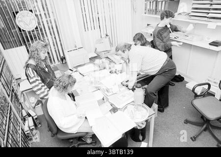 Five People Employment Office, Whizgle News, Dutch Desk, Niederlande, 1950 - 2000 am 25-01-1995. Die Abbildung zeigt diese Themen. Die Szene zeigt eine geschäftige Büroumgebung voller organisierter Chaos. In der Mitte ist ein großer Tisch mit Papieren, Ordnern und verschiedenen Büromaterialien überfüllt, was auf eine laufende Zusammenarbeit hindeutet. Mehrere Personen haben verschiedene Aufgaben: Zwei Personen, die am Tisch sitzen, überziehen Dokumente, während eine andere in der Nähe steht und aktiv schreibt oder tippt. Die Farbpalette ist überwiegend monochrom, wobei verschiedene Grauschattierungen entstehen Stockfoto