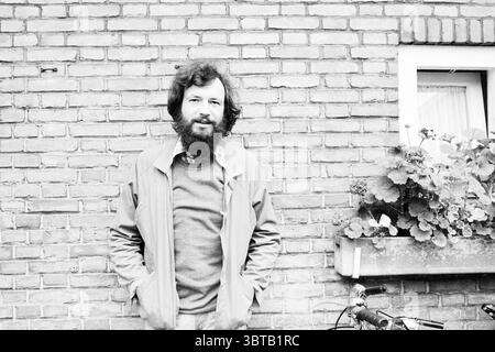 Porträt Ernst Oostenveld Komponist Musikporträts, Whizgle News, Dutch Desk, Niederlande, 1950 - 2000 am 06-10-1981. Diese Themen sind in der Abbildung dargestellt. In der Szene steht ein Mann selbstbewusst gegen eine strukturierte Ziegelwand. Seine Kleidung besteht aus einer hellen Jacke über einem Pullover, was ihm ein entspanntes, aber durchdachtes Aussehen verleiht. Sein langes, dunkles Haar und sein Vollbart tragen zu seinem unverwechselbaren Look bei und deuten auf ein kontemplatives Verhalten hin. Seitlich lehnt sich ein Fahrrad lässig an die Wand und verleiht dem Ambiente ein Gefühl des Alltags und der Bewegung. Der Rahmen des Fahrrads ist dunkel, Concept Stockfoto