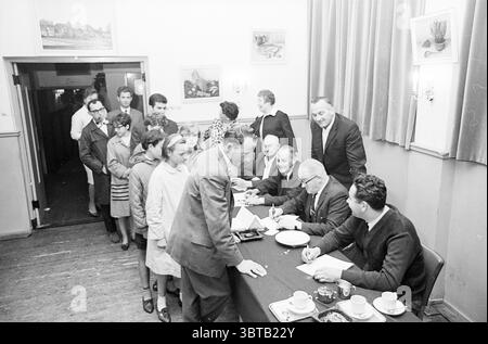 Anmeldungen Ringstiche Spiele und Spielzeug, Whizgle News, Dutch Desk, Niederlande, 1950 - 2000 am 02-08-1965. Diese Themen werden in der Abbildung angezeigt. Die Szene spielt in einem bescheidenen Innenraum, wahrscheinlich in einem Gemeindezentrum oder Saal. Es sind verschiedene Personengruppen anwesend, mit einer Mischung aus Erwachsenen und Kindern. Im Vordergrund ist ein langer Tisch von mehreren Männern besetzt, von denen einige Papiere schreiben oder unterschreiben. Sie tragen formelle Kleidung – Anzüge und Krawatten –, die auf ein bedeutendes Ereignis hindeutet. Der Tisch ist mit kleinen Tassen und Tassen geschmückt, die auf Erfrischungen hinweisen. Be Stockfoto